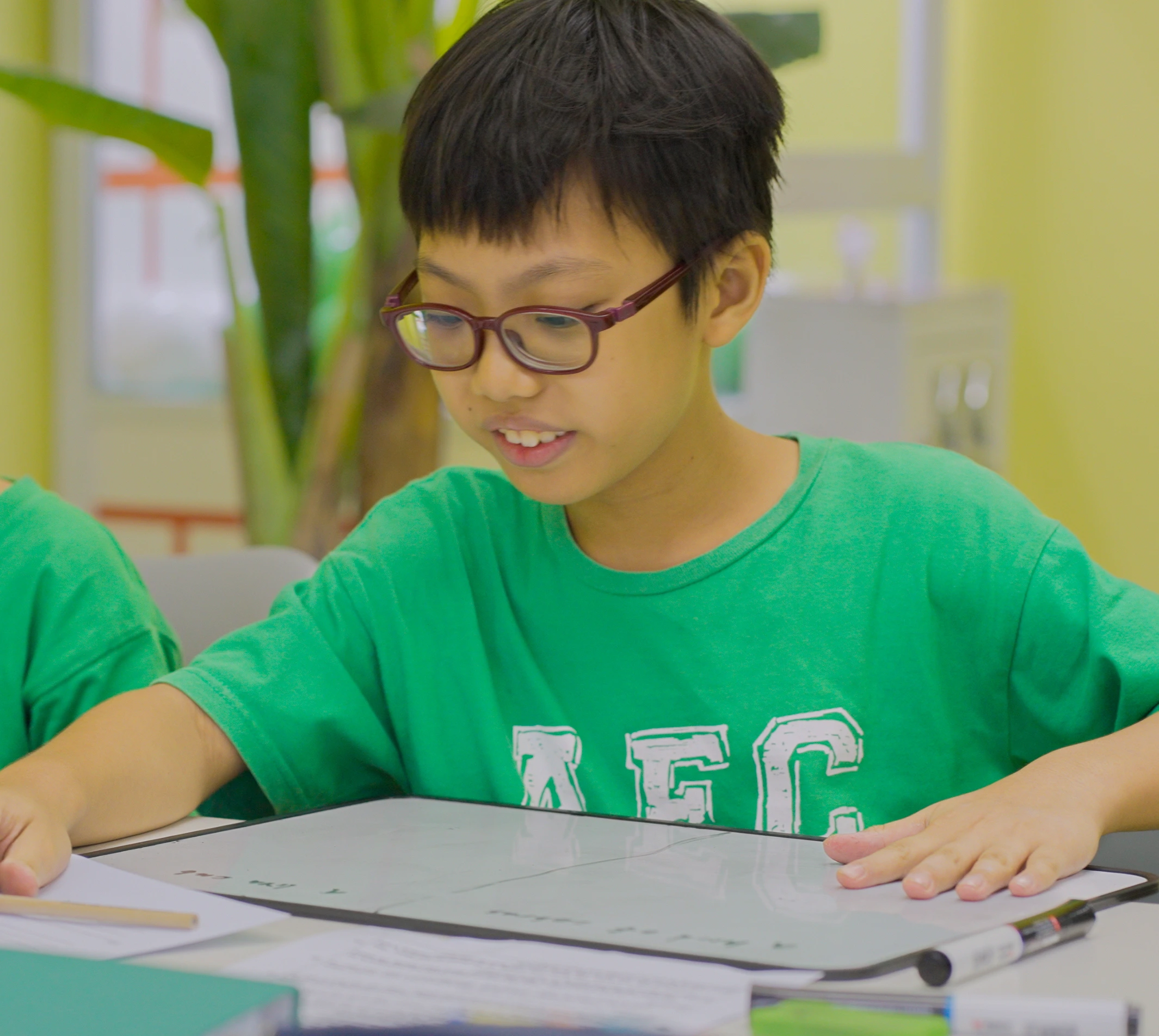 Young boy in glasses and a green shirt working intently on a whiteboard during an English lesson at Augustine's English Class.