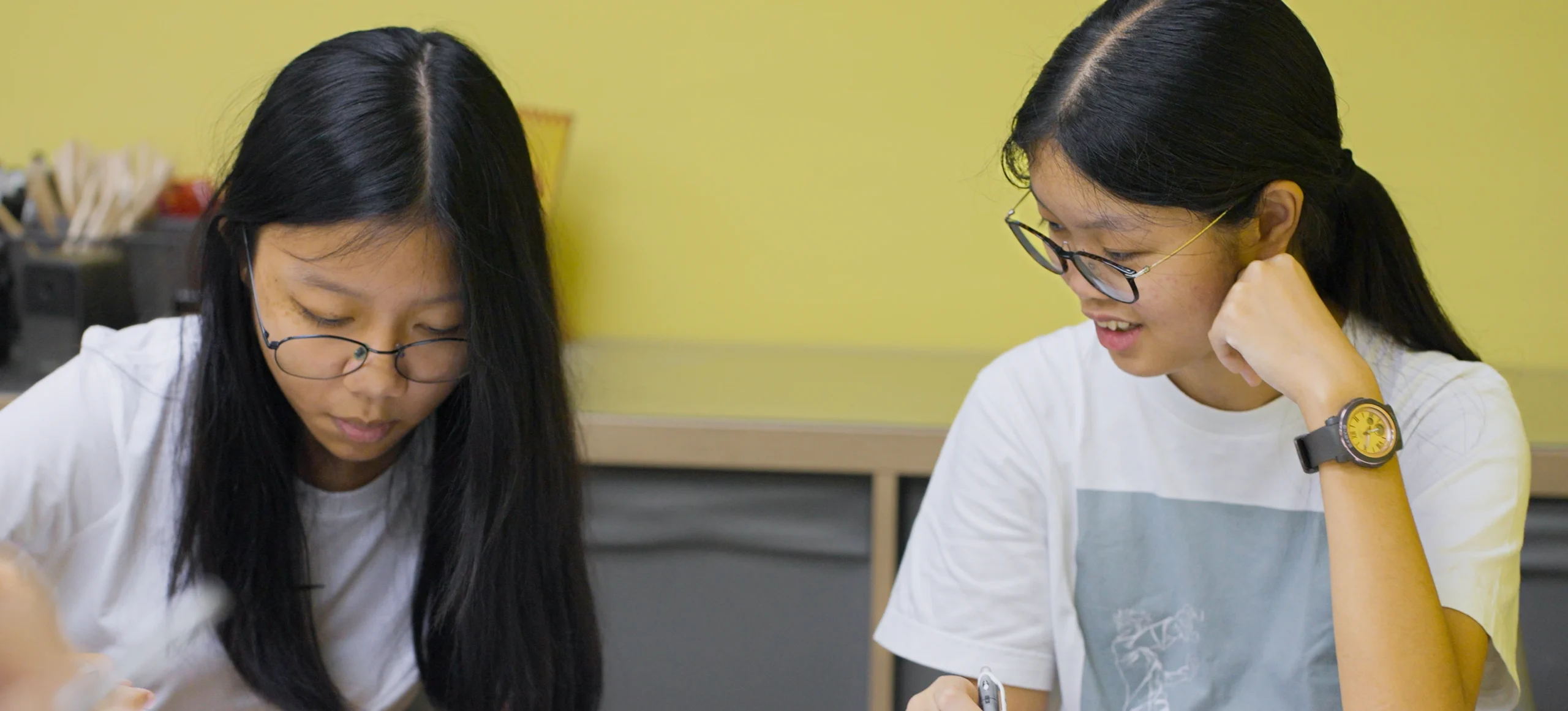 Two young Asian women with long black hair and glasses studying or collaborating at a table with a yellow background, one looking down intently, the other smiling slightly while doing an English writing test at the top English Tutorial centre in Singapore.