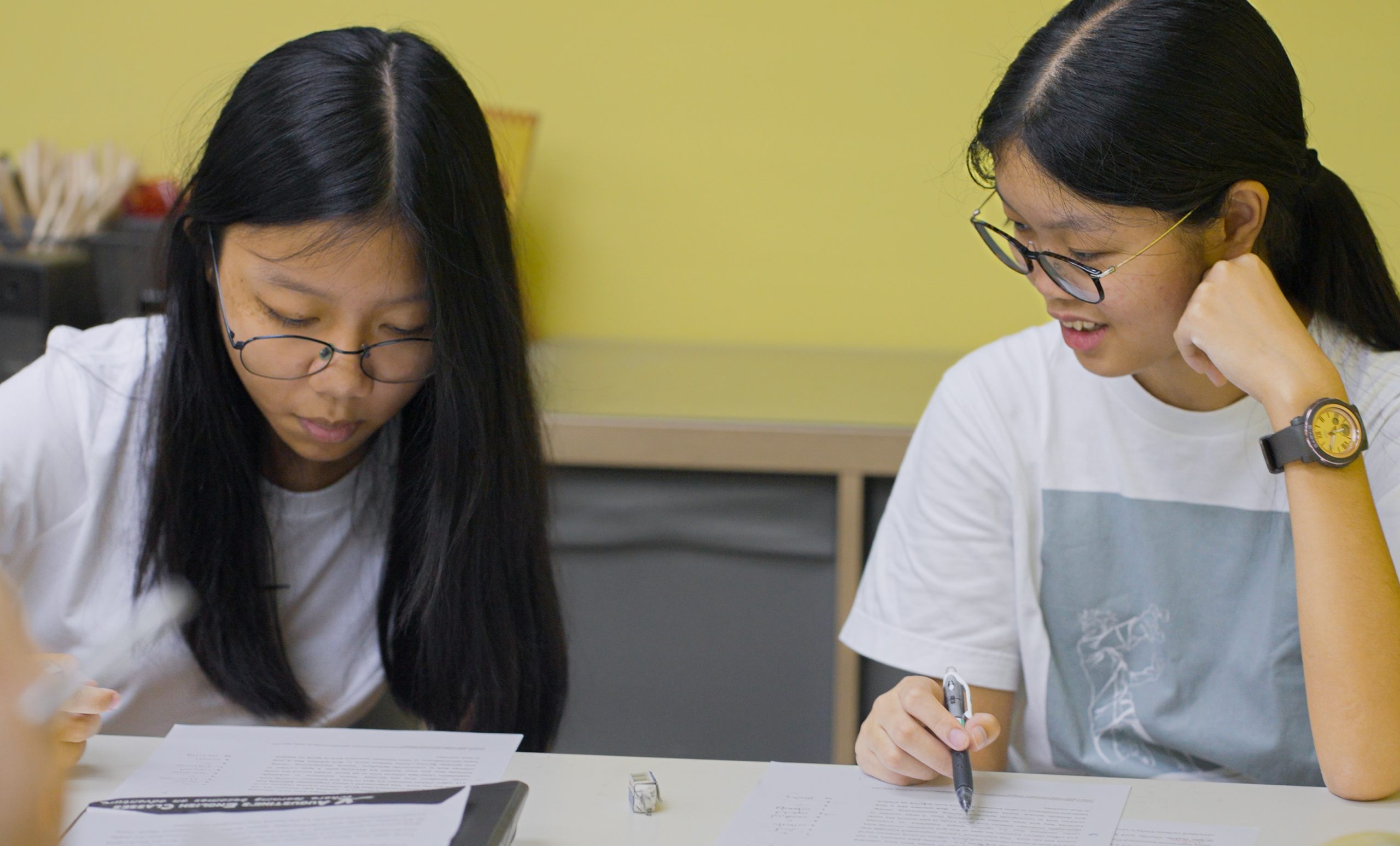 Two young Asian women intently studying English papers at a table at Augustine's English Centre, one with a pen in hand.
