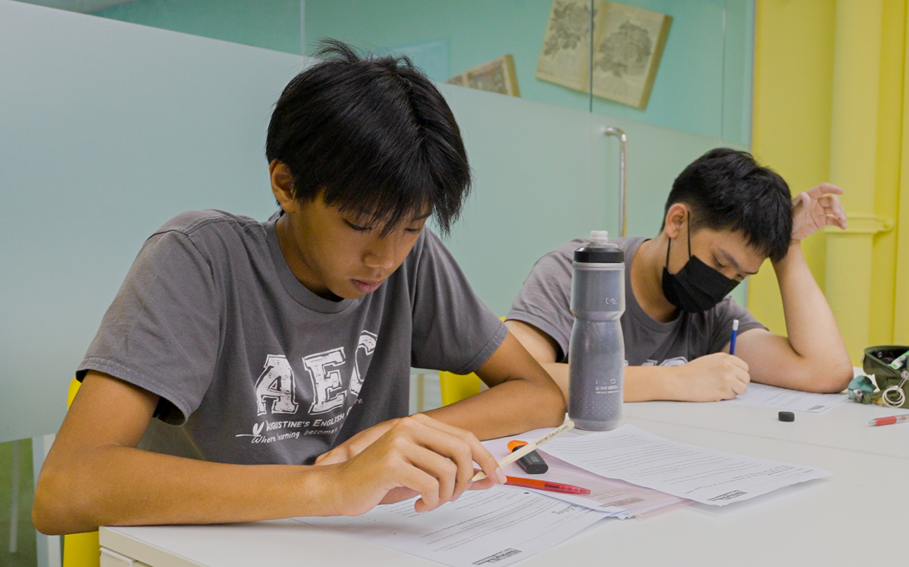 Two students diligently studying English at a white desk in a brightly lit classroom, one masked.