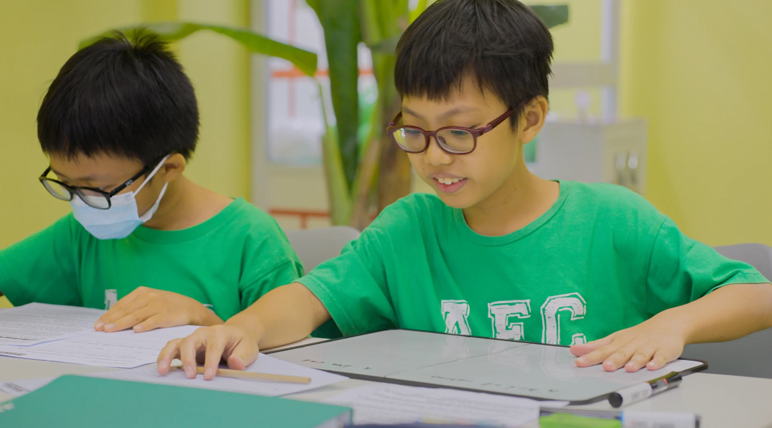 Two Asian boys in green shirts studying English at a desk in the Augustine's English Centre, one wearing glasses and a face mask, the other wearing glasses and looking at a small whiteboard.