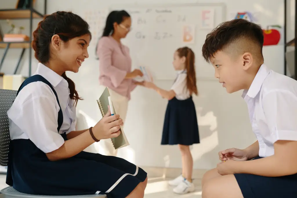  Two kids in the foreground reading from a notebook while a teacher hands something to another child in the background.
