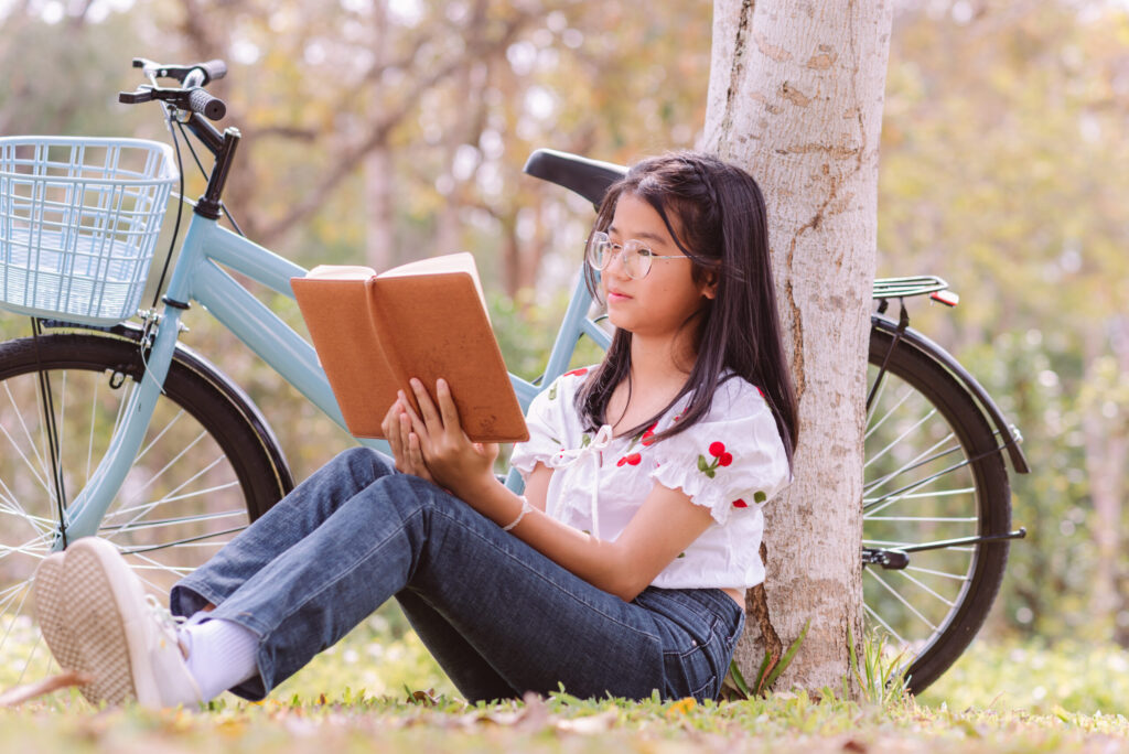 Secondary school student reading a book while sitting under a tree beside a bicycle, representing independent reading and critical thinking skills for secondary English learning.