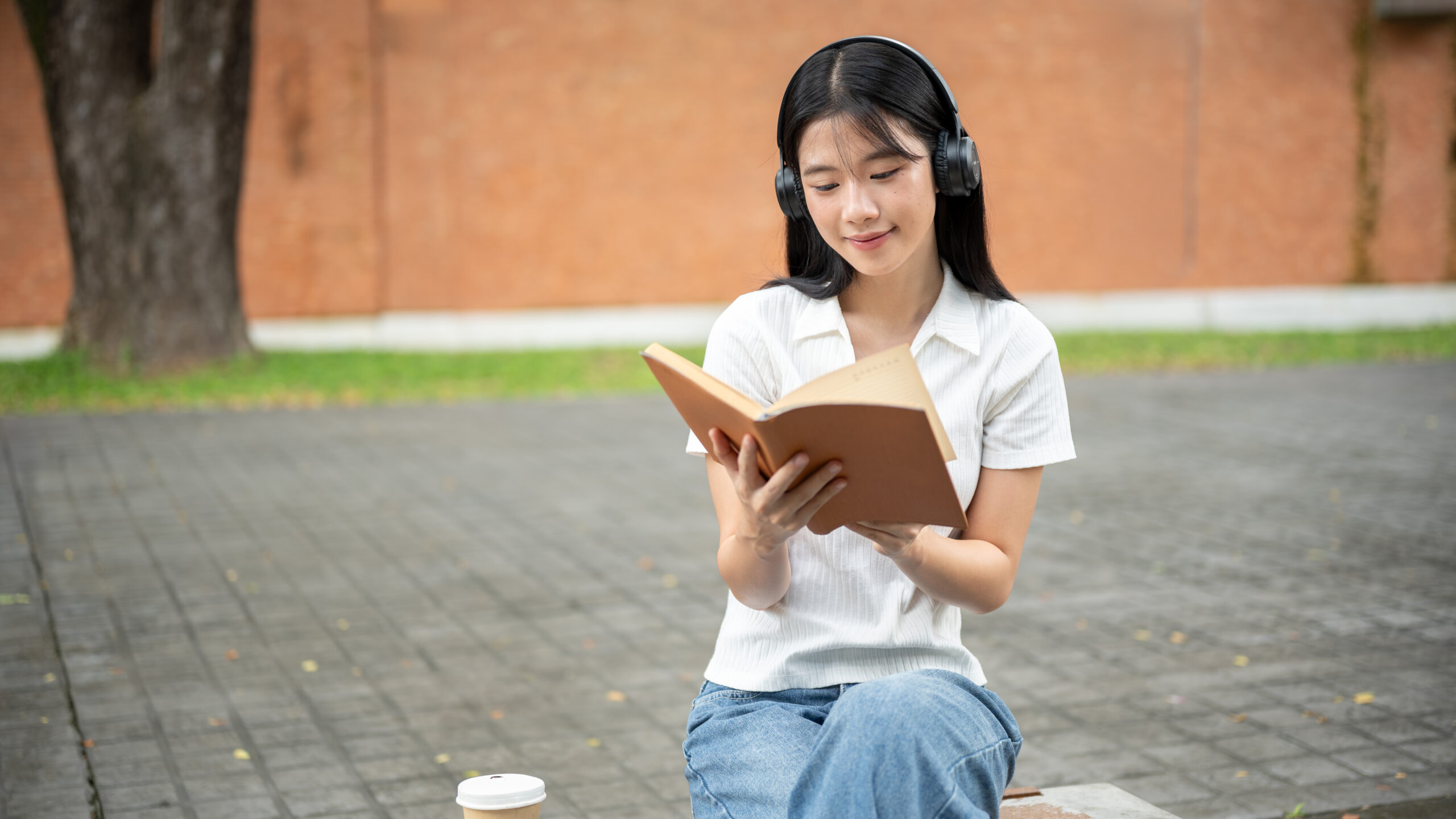 Secondary school student reading a book outdoors while wearing headphones, representing independent reading, critical thinking, and upper secondary English learning.